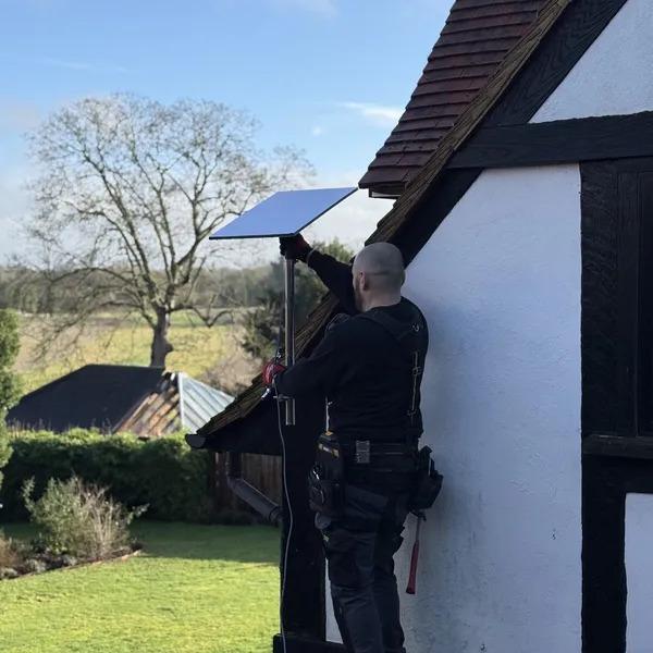 Engineer mounting a satellite dish on a tall pole against a timber framed house during a residential install