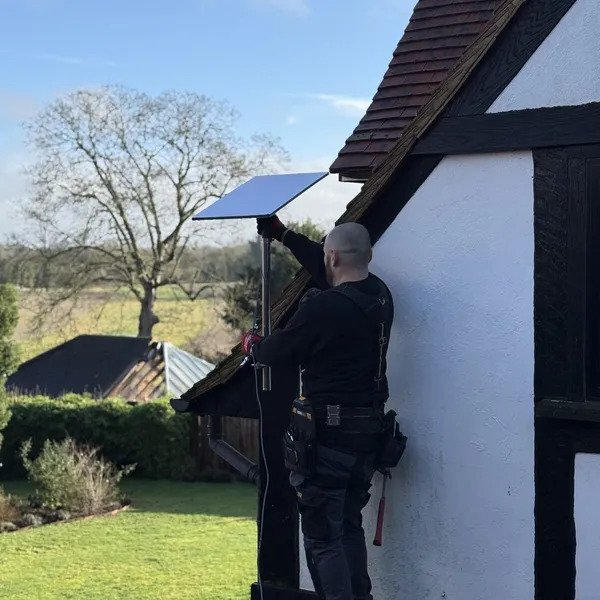 Engineer installing a Starlink dish on a tall pole mount against a timber framed house