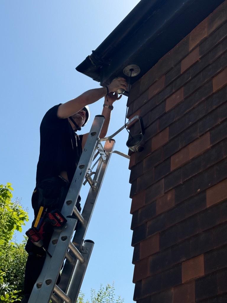 CCTV engineer in a safety helmet on a ladder fitting a dome camera under a brick house soffit