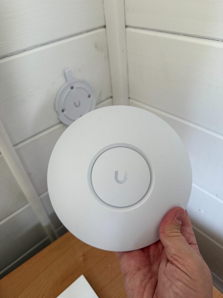 UniFi access point held in front of its wall mount on white timber cabin cladding inside a garden office